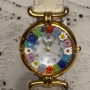 May include: A gold-tone wristwatch with a white dial featuring a ring of colorful millefiori-style floral glass beads around the perimeter. The watch has a textured off-white leather strap and gold-tone hands with the word Quartz printed on the face.