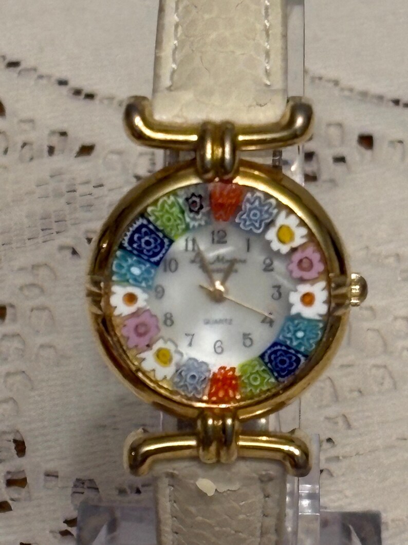 May include: A gold-tone wristwatch with a white dial featuring a ring of colorful millefiori-style floral glass beads around the perimeter. The watch has a textured off-white leather strap and gold-tone hands with the word Quartz printed on the face.