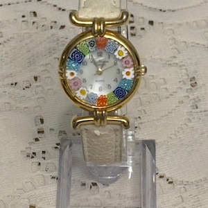 May include: A gold-tone wristwatch with a white textured leather strap. The round watch face features a white dial with black numerals and a colorful millefiori glass border containing floral patterns in blue, orange, pink, and white.
