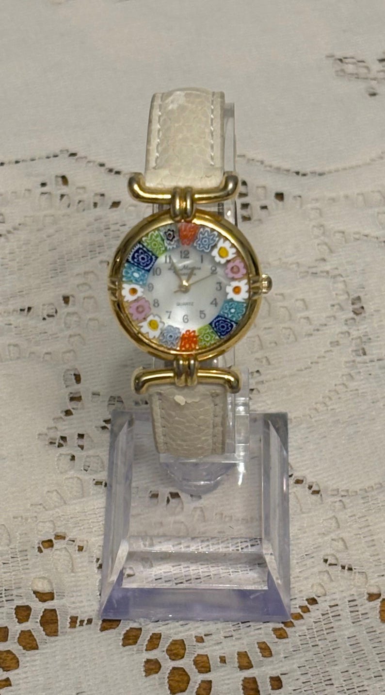 May include: A gold-tone wristwatch with a white textured leather strap. The round watch face features a white dial with black numerals and a colorful millefiori glass border containing floral patterns in blue, orange, pink, and white.