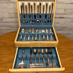 May include: A wooden flatware chest with a hinged lid and a pull-out drawer, featuring a blue interior lining. The set includes various silver-toned metal knives, forks, and spoons arranged in designated slots, including a large serving spoon.