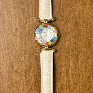 May include: A wristwatch with a gold-tone circular case and a white textured leather strap. The watch face features a colorful floral pattern around the perimeter, black numerals, and thin gold-tone hands. The buckle is gold-tone metal.