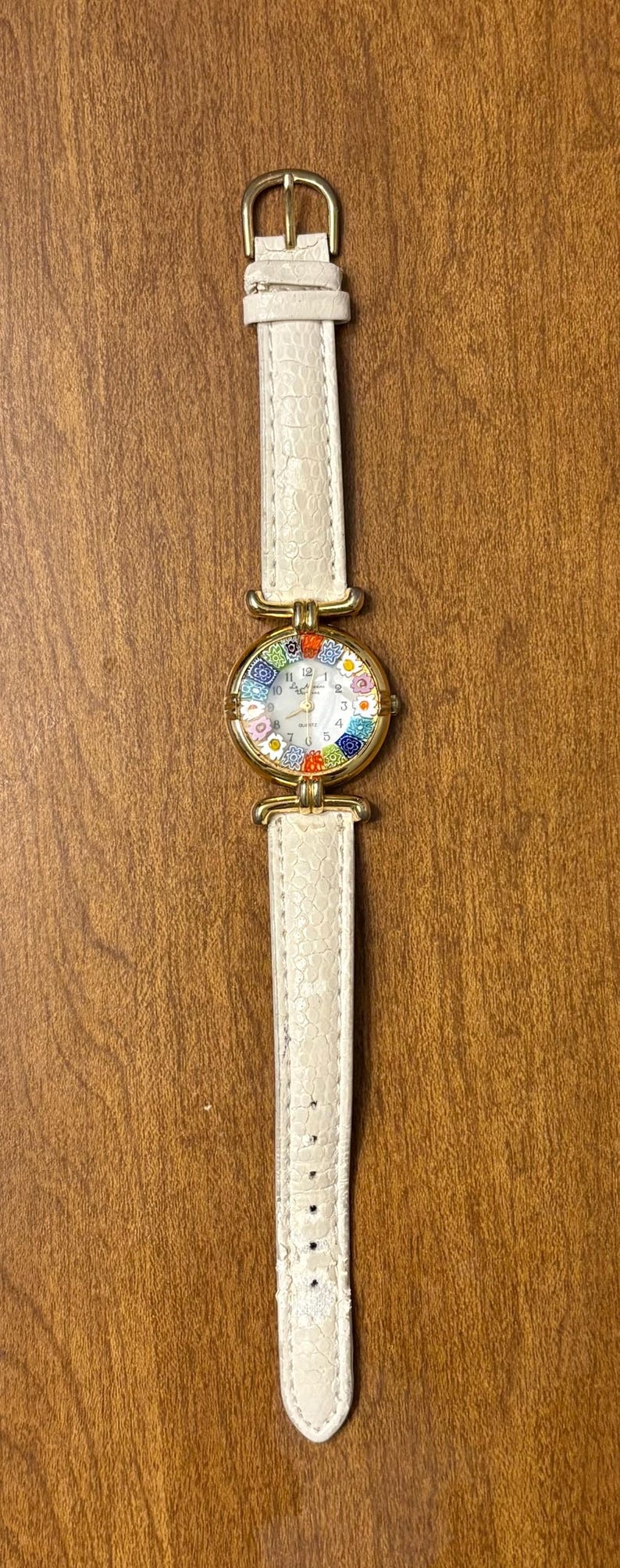 May include: A wristwatch with a gold-tone circular case and a white textured leather strap. The watch face features a colorful floral pattern around the perimeter, black numerals, and thin gold-tone hands. The buckle is gold-tone metal.