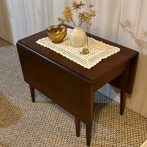 May include: A dark brown wooden drop-leaf table featuring a cream-colored crocheted doily, an amber glass bowl, a small white ceramic vase with dried flowers, and a tiny brass animal figurine placed on top of the table surface.