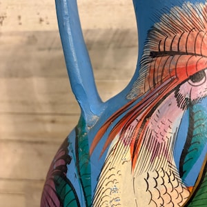 May include: A hand-painted ceramic pitcher with a vibrant bird design. The pitcher is primarily blue with a detailed illustration of a bird in shades of red, white, and green. The handle is a solid blue.