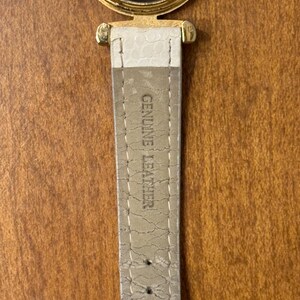May include: A gold-tone watch case with a stainless steel back featuring the text QUARTZ and MOVEMENT. Attached is a light beige watch strap with a textured surface and the embossed text GENUINE LEATHER, showing visible signs of wear and cracking.