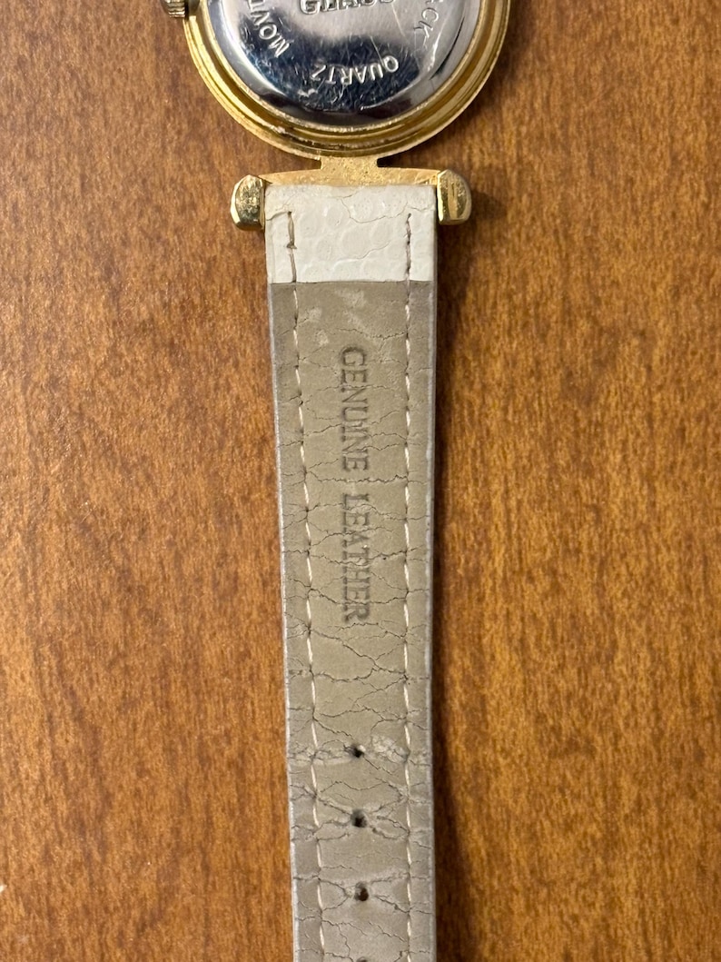 May include: A gold-tone watch case with a stainless steel back featuring the text QUARTZ and MOVEMENT. Attached is a light beige watch strap with a textured surface and the embossed text GENUINE LEATHER, showing visible signs of wear and cracking.