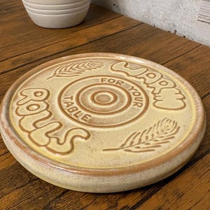 May include: A round, ceramic trivet with a wheat and concentric circle design. The trivet is tan with brown accents and features the words "Buns for your table". A cream-colored vase is in the background.