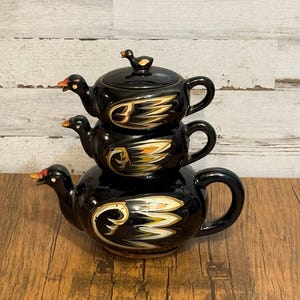 May include: A stacked black ceramic teapot set with a duck theme. The set includes three teapots, each with a duck spout and handle. The teapots are decorated with gold and white wing designs.