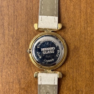 May include: A wristwatch with a gold-tone circular case and a white textured strap. The back of the watch case is silver-tone stainless steel and features the text MURANO GLASS, STAINLESS STEEL BACK, and QUARTZ MOVT. The strap is marked GENUINE.