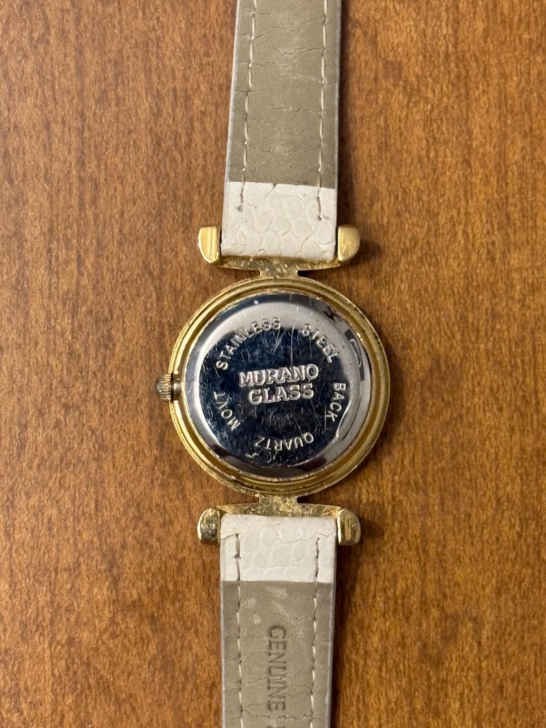 May include: A wristwatch with a gold-tone circular case and a white textured strap. The back of the watch case is silver-tone stainless steel and features the text MURANO GLASS, STAINLESS STEEL BACK, and QUARTZ MOVT. The strap is marked GENUINE.