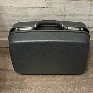 May include: A dark gray Samsonite suitcase with a black handle and silver accents. The suitcase has a rectangular shape with rounded corners and a textured surface. The handle is centered on the top.
