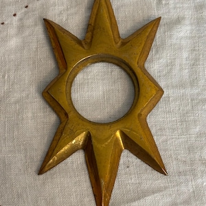 May include: A gold-colored metal star-shaped object with six pointed rays and a circular opening in the center. The surface has a smooth, metallic finish with subtle variations in tone, creating a reflective appearance across the points.