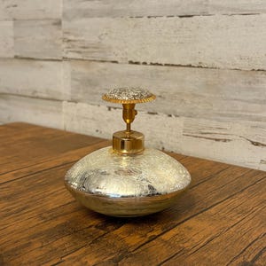 May include: A decorative perfume bottle with a silver, textured body and a gold-colored top. The bottle has a round shape and a small, decorative stopper. The stopper is gold-colored with a textured, silver top.