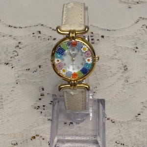 May include: A wristwatch with a gold-tone circular case featuring a colorful millefiori glass bezel with floral patterns. The watch has a white dial with black numerals and a textured off-white leather strap with gold-tone metal hardware connectors.