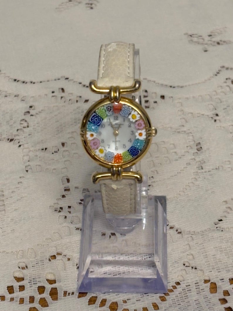 May include: A wristwatch with a gold-tone circular case featuring a colorful millefiori glass bezel with floral patterns. The watch has a white dial with black numerals and a textured off-white leather strap with gold-tone metal hardware connectors.