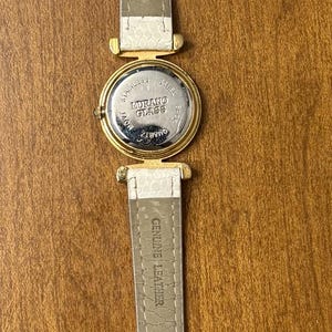 May include: A wristwatch with a white textured leather strap and a gold-tone metal case. The back of the watch case is stainless steel and features the text Murano Glass, Quartz, and Movt. The strap is marked Genuine Leather on the inner side.