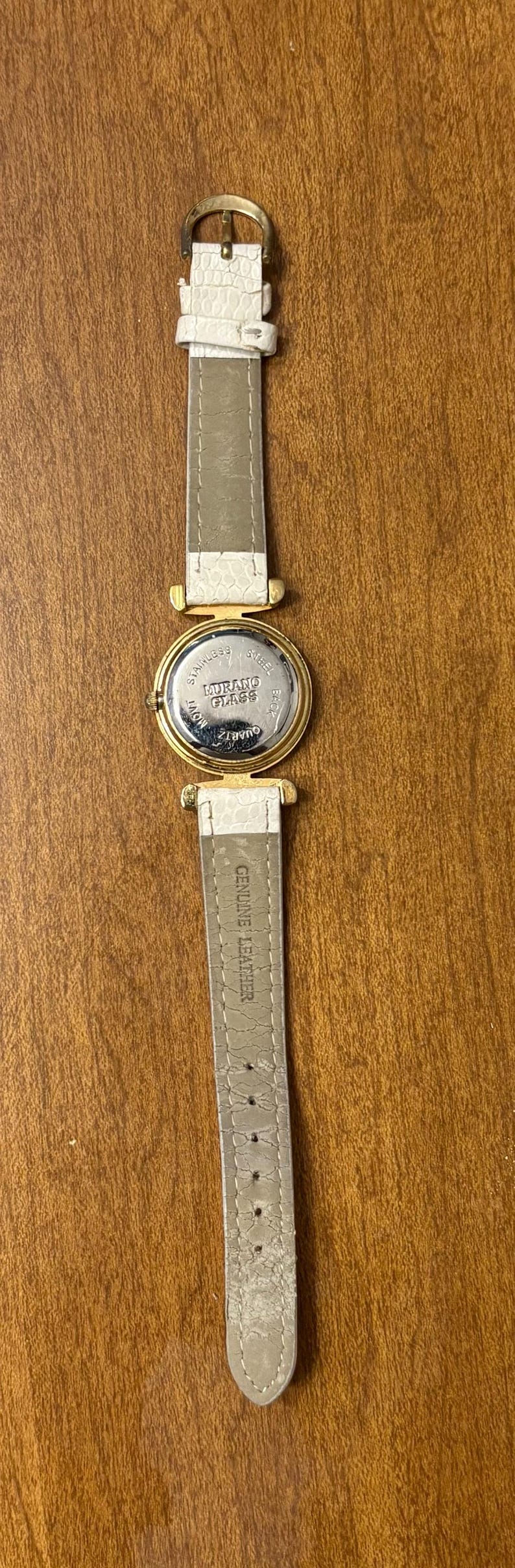 May include: A wristwatch with a white textured leather strap and a gold-tone metal case. The back of the watch case is stainless steel and features the text Murano Glass, Quartz, and Movt. The strap is marked Genuine Leather on the inner side.