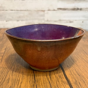 May include: A handmade ceramic bowl with a deep purple interior and a gradient exterior transitioning from orange to brown. The bowl has a slightly irregular shape and a rustic, earthy aesthetic.