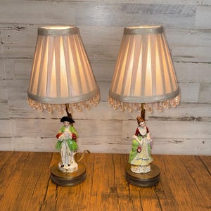 May include: Pair of vintage porcelain table lamps. Each lamp features a pleated lampshade with beaded trim, and a base with a figurine of a man and woman in period clothing. The lamps are on a wooden surface.