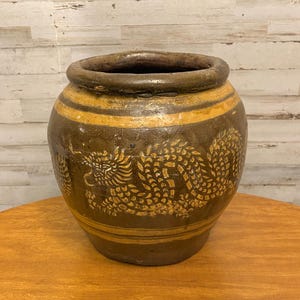 May include: A large, round, dark brown ceramic jar featuring a wide opening and a thick rim. The exterior is decorated with a prominent, light-colored, etched dragon motif encircling the body, accented by horizontal bands near the top and base.