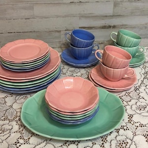 May include: A collection of pastel-colored ceramic dishware, including plates, saucers, and teacups. The set features pink, blue, and green pieces with a scalloped edge design. The arrangement suggests a tea party or dining setting.