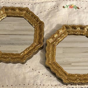 May include: Two octagonal mirrors featuring ornate, textured gold-toned frames with scalloped edges. The reflective glass surfaces are clear and set within the detailed, raised metallic borders that exhibit a repeating pattern.