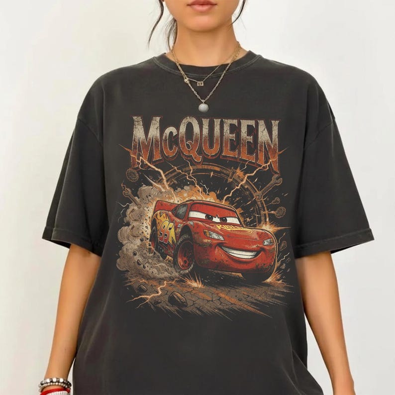 Peut inclure: Un t-shirt gris anthracite &agrave; manches courtes avec une impression graphique d'une voiture de course rouge portant le num&eacute;ro 95 sur le c&ocirc;t&eacute;. Le texte MCQUEEN est imprim&eacute; en grandes lettres majuscules us&eacute;es au-dessus de la voiture entour&eacute;e de poussi&egrave;re.