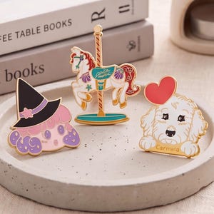 Op de afbeelding: Drie emaille pins met een personage met een zwarte heksenhoed en roze haar, een wit carrouselpaard met gouden accenten, en een witte hond met een rood hart boven het hoofd en de naam Carmela onderaan de rand gedrukt.