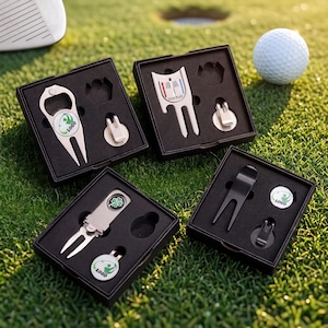 Könnte beinhalten: Vier Golf-Tool-Sets in schwarzen Boxen, jeweils mit einem Pitchgabel, einem Ballmarker und einem magnetischen Hutclip. Ein Set hat einen Flaschenöffner. Die Werkzeuge sind silber und schwarz, mit grünen und weißen Logos. Ein Golfball ist im Hintergrund.