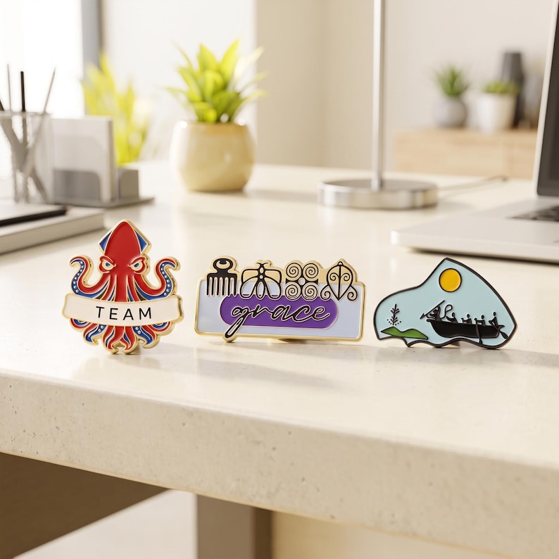 May include: Three enamel pins displayed on a desk. The first is a red and blue octopus with the word TEAM. The second is a rectangular pin with purple accents, symbols, and the word grace. The third is a light blue landscape with a boat and sun.