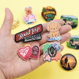 May include: A collection of colorful, decorative pins and magnets. Designs include a poodle, a road trip sign, a heart-shaped box of chocolates, and a puzzle piece with the letter "B". The pins have various texts, including "Road Trip" and "Almond Fest 2026".
