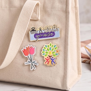 May include: A beige canvas tote bag featuring three enamel pins: a rectangular pin with the word grace and four symbols, a bouquet of three pink and orange tulips, and a colorful tree design with three stylized figures at the base.