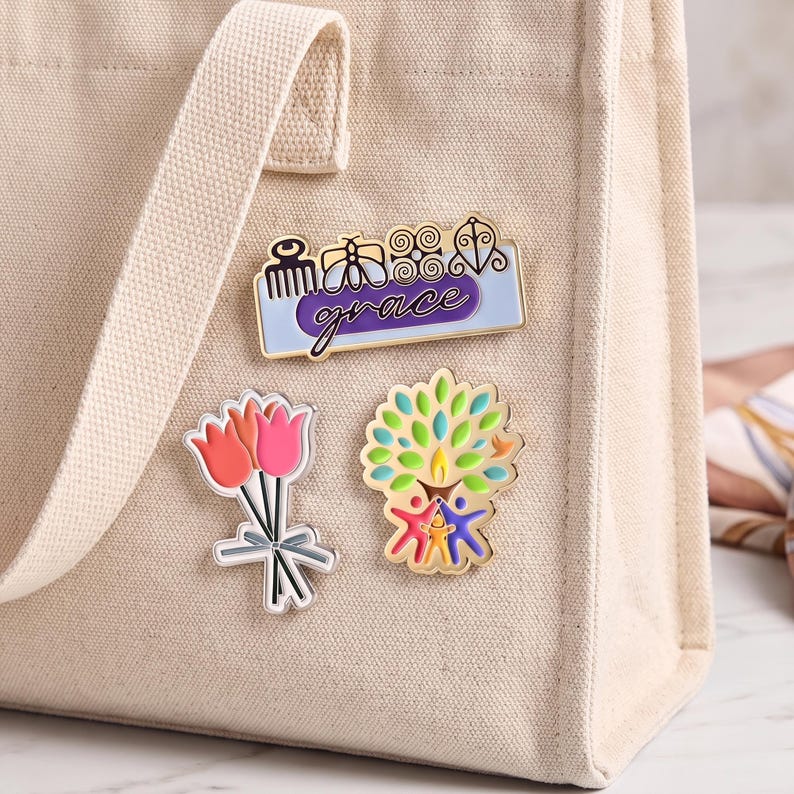 May include: A beige canvas tote bag featuring three enamel pins: a rectangular pin with the word grace and four symbols, a bouquet of three pink and orange tulips, and a colorful tree design with three stylized figures at the base.