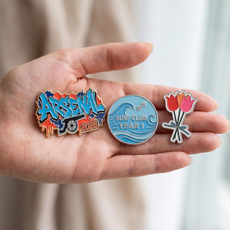 May include: Three enamel pins: one irregular shape with blue graffiti text reading ARSENAL and 2026 over a cannon, one circular light blue pin with wave graphics and text reading SURF CLUB YEAR 1, and one pin shaped like a bouquet of three tulips.