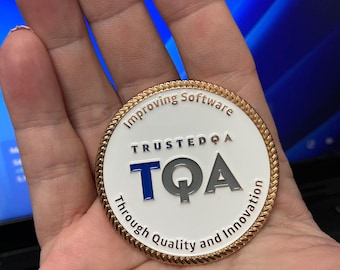 May include: A round metal coin with a white enamel center and a gold-toned rope-textured border. The text "TRUSTEDQA" appears above the large letters "TQA" in blue and grey, with "Improving Software Through Quality and Innovation" around the edge.