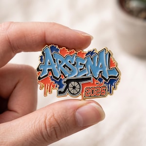 May include: A metal enamel pin featuring the word ARSENAL in blue graffiti-style lettering with black outlines. The design includes a black cannon icon and the orange text 2026, set against a background of orange and blue paint drip patterns.