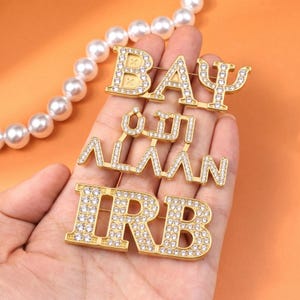 Puede incluir: Broches dorados que deletrean "BAY", "ALAAAN" en escritura árabe e "IRB", todos incrustados con pedrería transparente. Un collar de perlas está en el fondo. Los broches se sostienen en una mano sobre un fondo naranja.