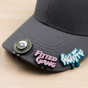 Puede incluir: Una gorra de béisbol gris carbón con tres pines metálicos sujetos a la visera. Los pines incluyen un diseño circular verde y plateado con el texto Gotta Catch Em All, un pin rosa y negro que dice Fitted Gang y un pin de color verde azulado con purpurina.