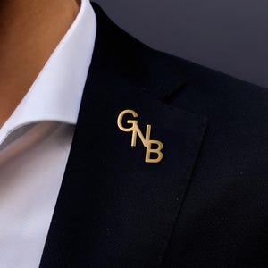 Op de afbeelding: Een goudkleurige reversspeld met de letters "GNB" in een gestileerd lettertype. De speld is bevestigd aan een zwart colbert, met daaronder een wit overhemd. Een stijlvol accessoire.
