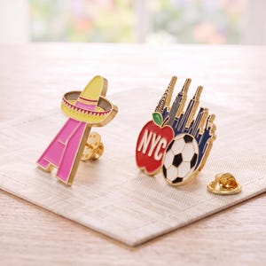 May include: Two metal enamel pins. One pin is a pink letter A topped with a yellow sombrero. The second pin features a red apple with the text NYC, a black and white soccer ball, and a dark blue silhouette of the New York City skyline.