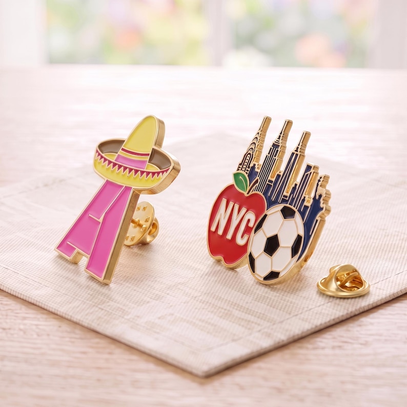 May include: Two metal enamel pins. One pin is a pink letter A topped with a yellow sombrero. The second pin features a red apple with the text NYC, a black and white soccer ball, and a dark blue silhouette of the New York City skyline.