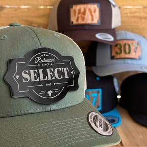 Könnte beinhalten: Eine Auswahl an Trucker-Caps in verschiedenen Farben, darunter Olivgrün, Braun und Grau. Jede Kappe hat ein einzigartiges Patch mit Text oder einer Zahl. Die olivgrüne Kappe hat ein schwarzes Patch mit der Aufschrift "Restaurant SELECT since 1982".