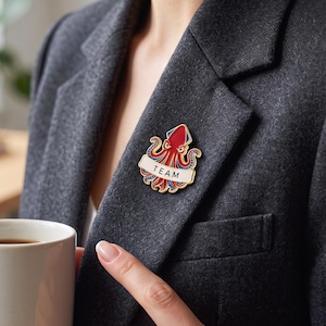 May include: A gold-toned enamel pin featuring a stylized red squid with blue accents and a white banner across the front that displays the word TEAM in black capital letters. The pin is attached to the lapel of a dark gray textured blazer.
