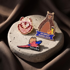 May include: Three enamel pins: one featuring a red glittery cartoon character with a wide grin, one shaped like a brown bear holding a flashlight with the text UCR WOMEN'S RESOURCE CENTER, and one depicting a colorful parrot with the text binske.