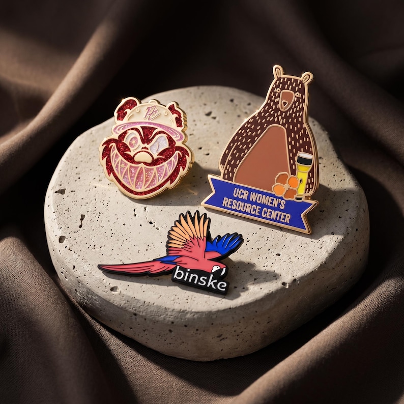 May include: Three enamel pins: one featuring a red glittery cartoon character with a wide grin, one shaped like a brown bear holding a flashlight with the text UCR WOMEN'S RESOURCE CENTER, and one depicting a colorful parrot with the text binske.