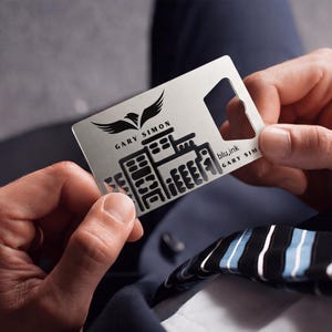 May include: A silver metal multi-tool with a bottle opener and a cityscape design. The tool features the text "GARY SIMON" and "blu.ink". An eagle graphic is above the name. The tool is held in two hands.