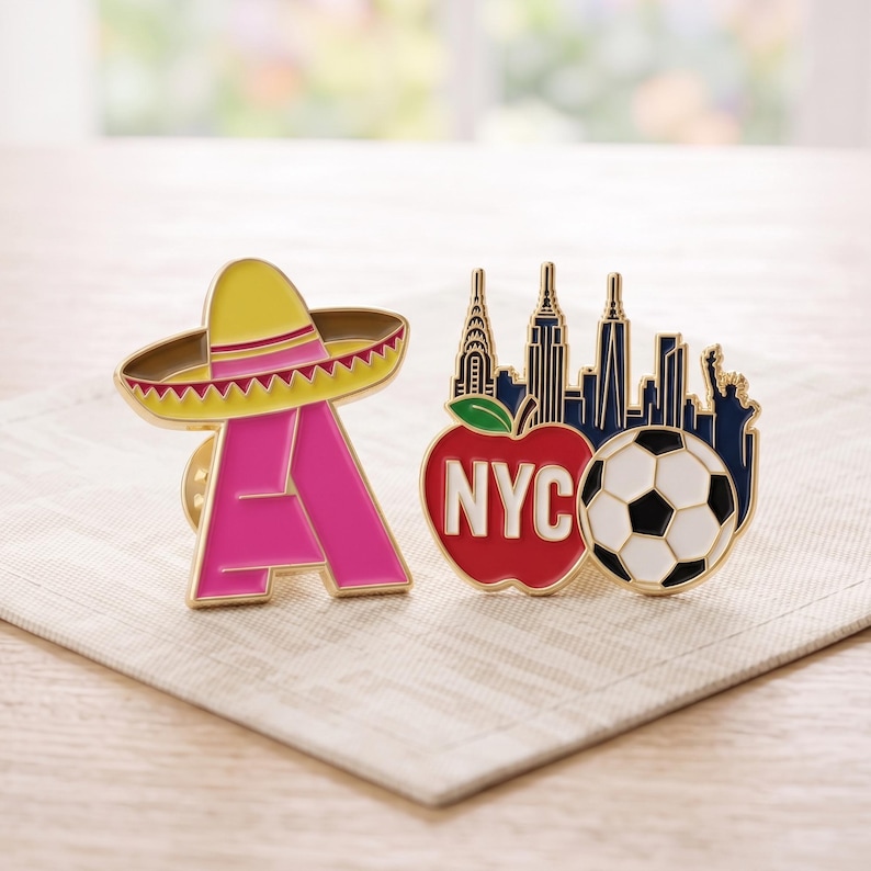 May include: Two enamel pins. One is a bright pink letter A topped with a yellow sombrero. The other features a dark blue New York City skyline silhouette, a red apple with the white text NYC, and a black and white soccer ball graphic.