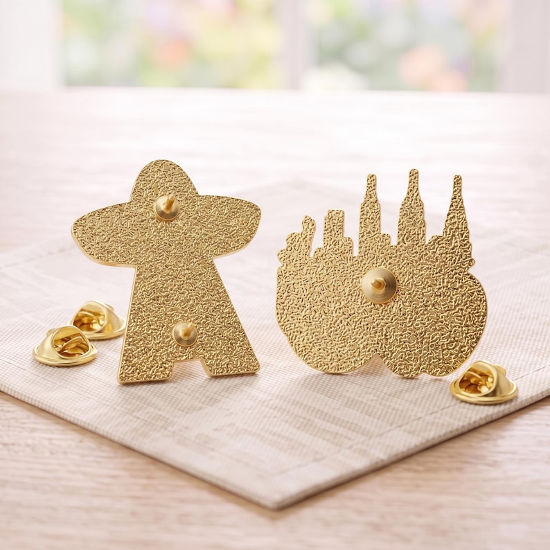 May include: Two gold-toned metal enamel pins shown from the back, featuring a textured surface. One pin is shaped like a game piece, and the other depicts a city skyline. Each pin includes two metal clutch backings with small circular fasteners.