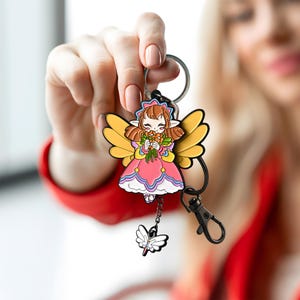 Könnte beinhalten: Ein farbenfroher Schlüsselanhänger mit einer Cartoon-Fee mit gelben Flügeln, die Blumen hält und ein rosa Kleid trägt. Der Schlüsselanhänger hat einen silbernen Ring und einen schwarzen Verschluss mit einem baumelnden Schmetterlingsanhänger.